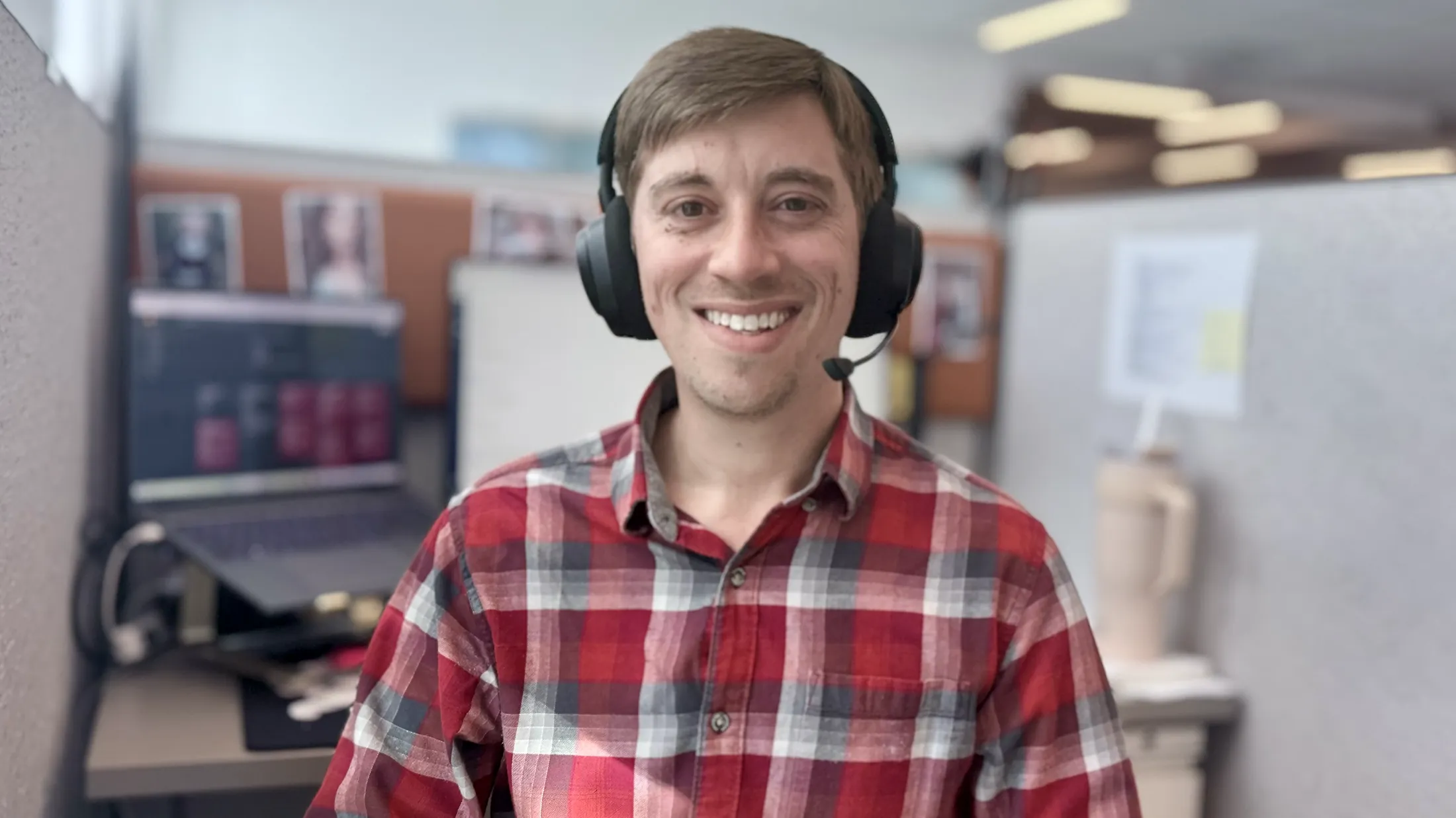 Smiling man wearing a red plaid shirt and black headset in an office cubicle.
