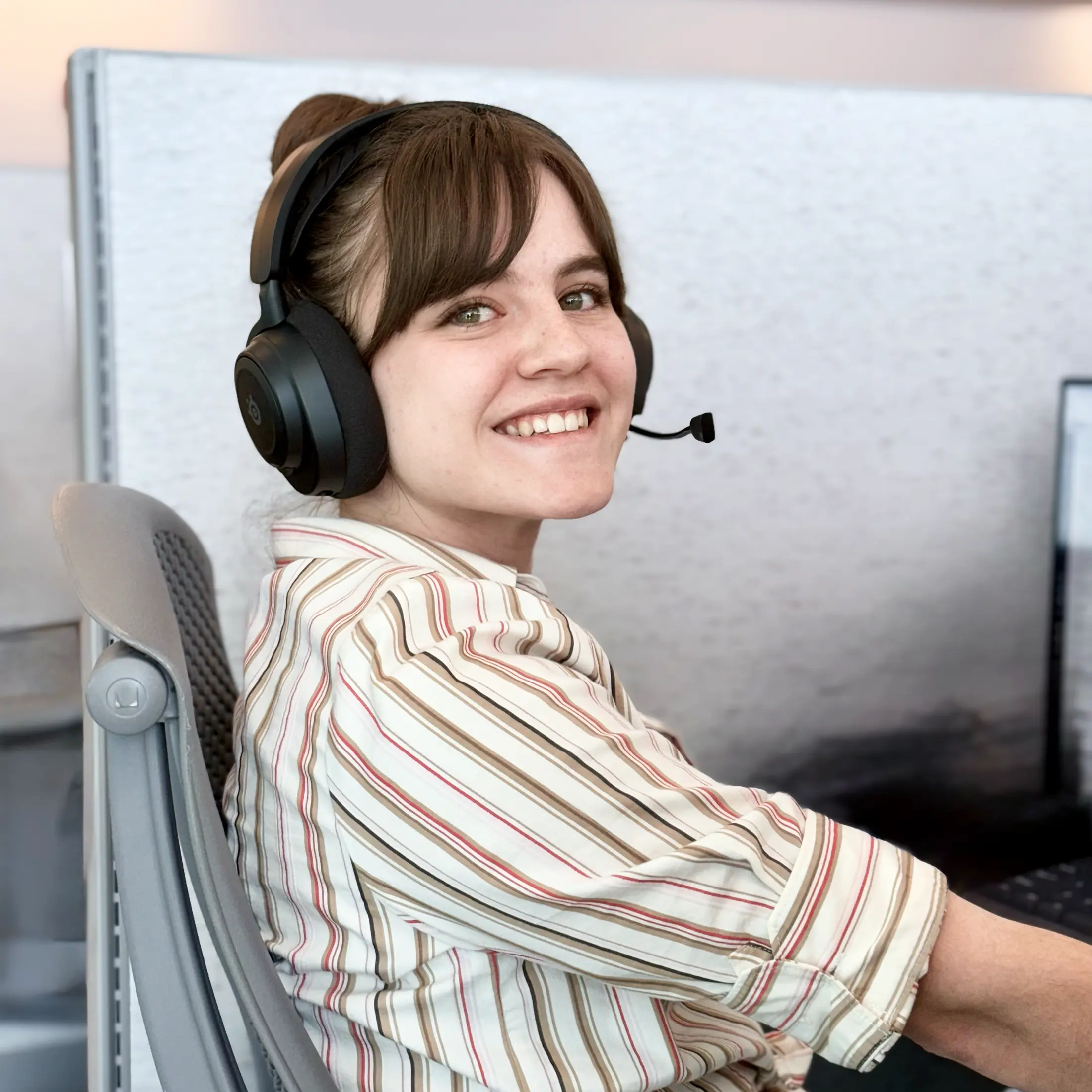 Smiling woman with brown hair wearing black headset and striped shirt, sitting in office chair at a computer.