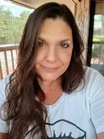 Woman with long brown hair wearing a white shirt, sitting on a porch with a wooden railing and trees in the background.