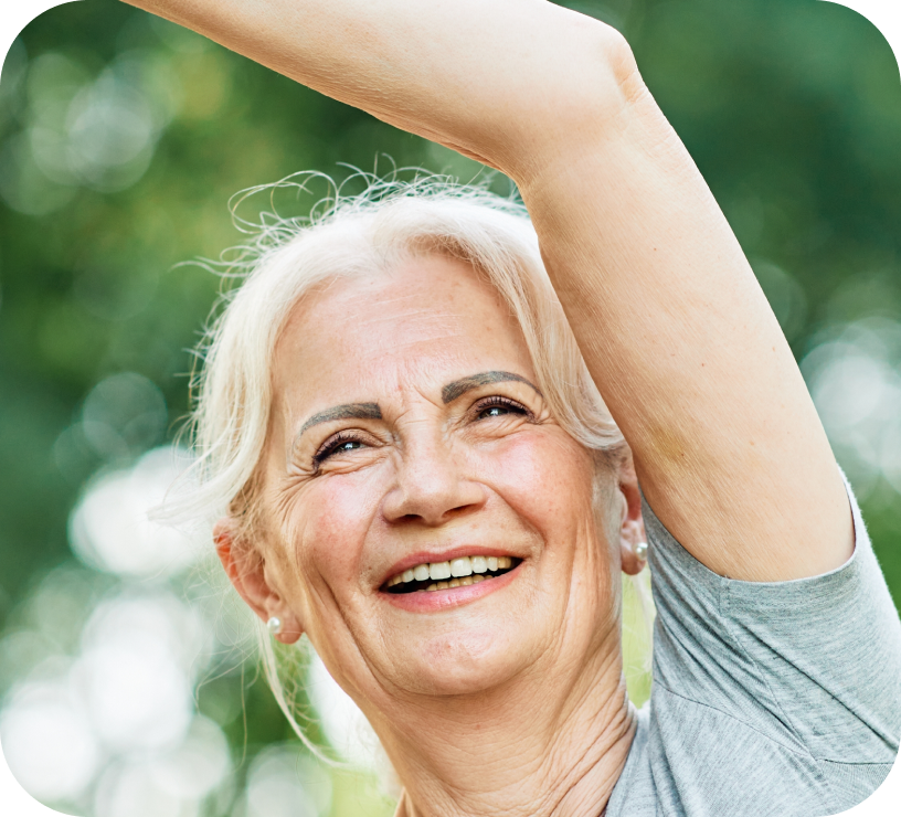 Smiling elderly woman with white hair raising her arm outdoors with blurred green foliage background.