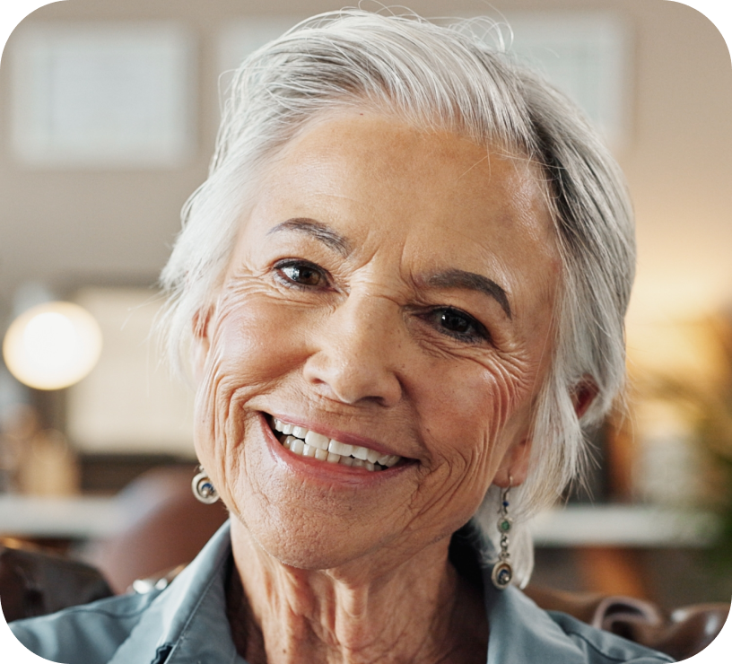 Smiling elderly woman with short gray hair and earrings in a softly lit indoor setting.