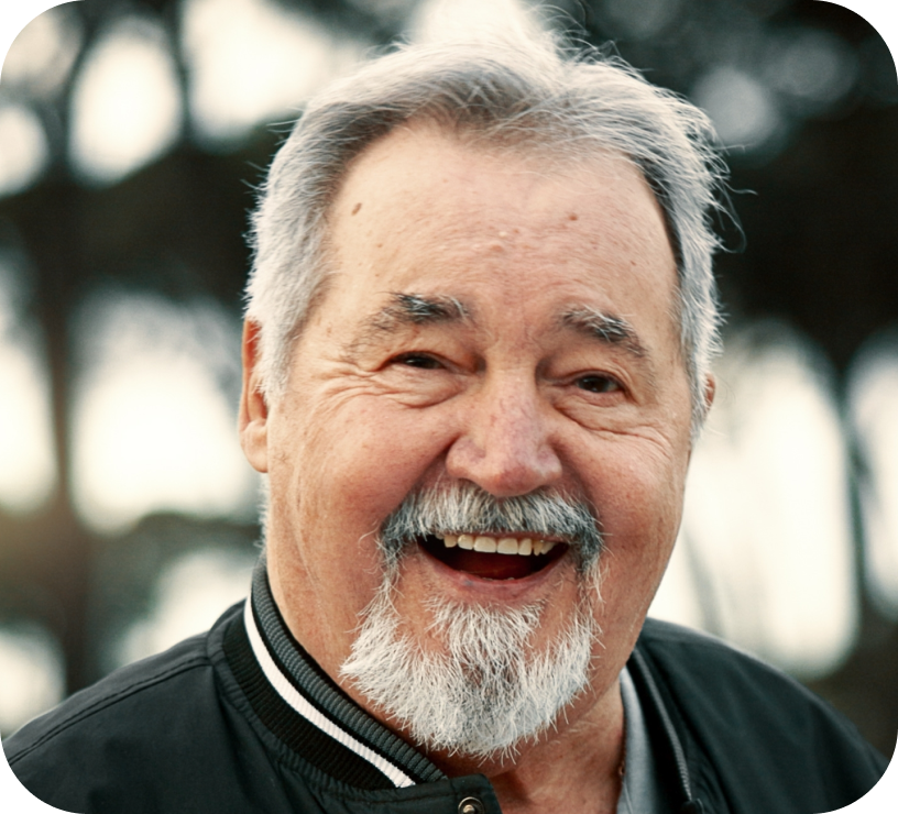Smiling elderly man with gray hair, beard, and mustache wearing a black jacket outdoors.