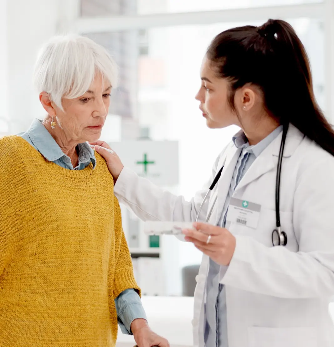 Female doctor with stethoscope comforting and speaking to an elderly woman in a mustard sweater.