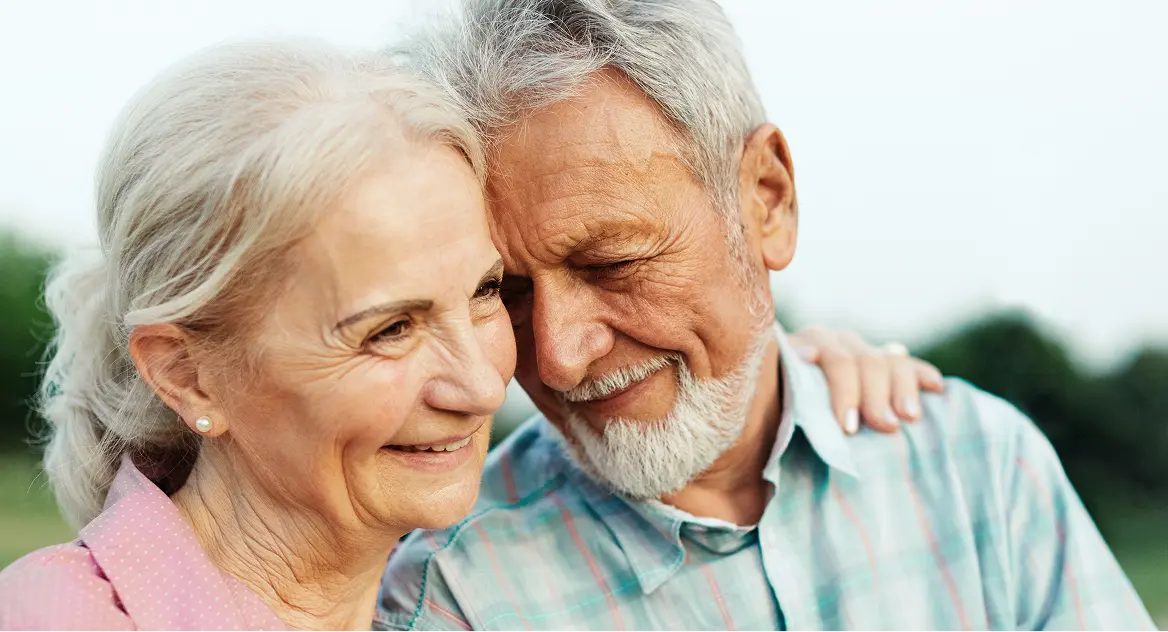 Smiling elderly couple embracing outdoors with the woman’s hand resting on the man’s shoulder.