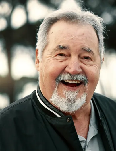Smiling elderly man with gray hair and beard wearing a black jacket outdoors.