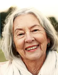 Smiling elderly woman with gray hair wearing a light-colored scarf outdoors.