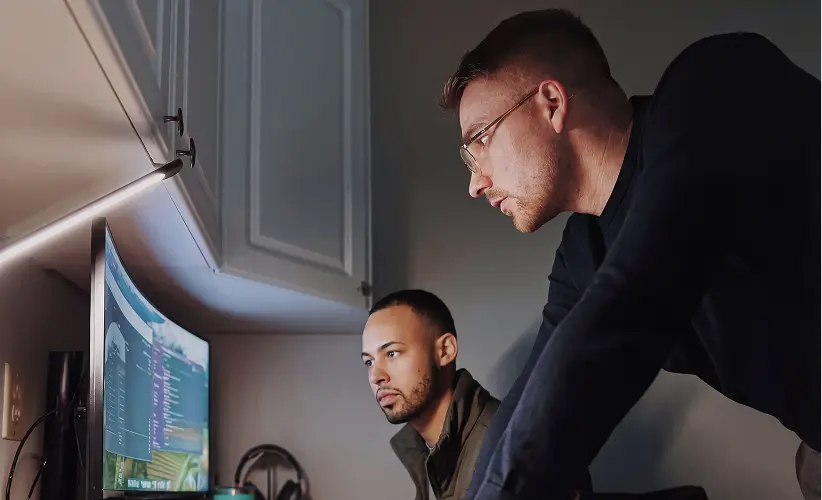 Two men focused on a curved computer monitor displaying code and data in a dimly lit room.