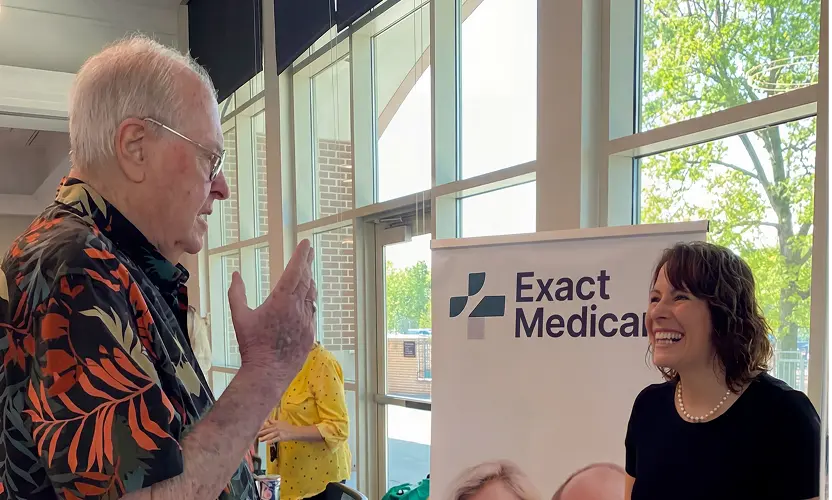 An elderly man speaking with a smiling woman standing beside an Exact Medicare banner in a bright room with large windows.