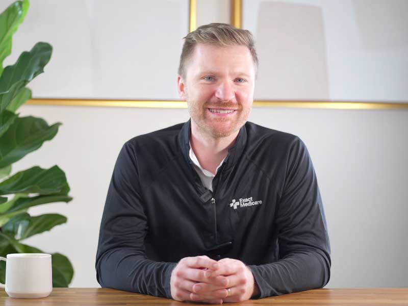 Smiling man in a black Exact Medicare jacket sitting at a wooden table with hands clasped, a plant and a white mug on the table.