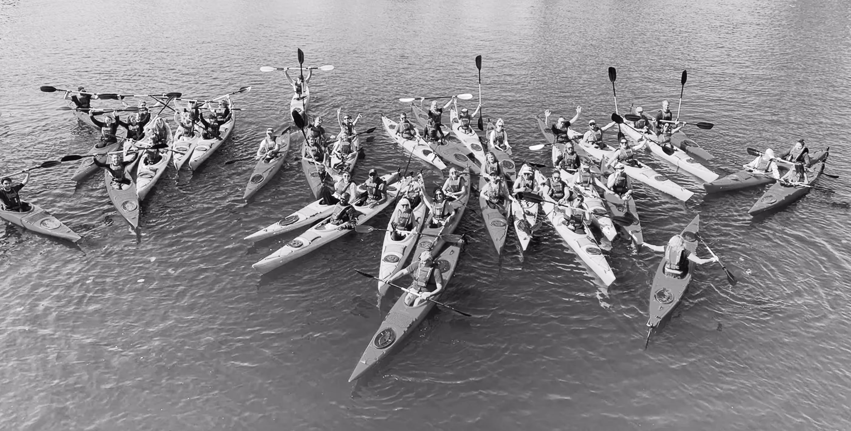 Group of people in kayaks on calm water, some raising paddles and waving.