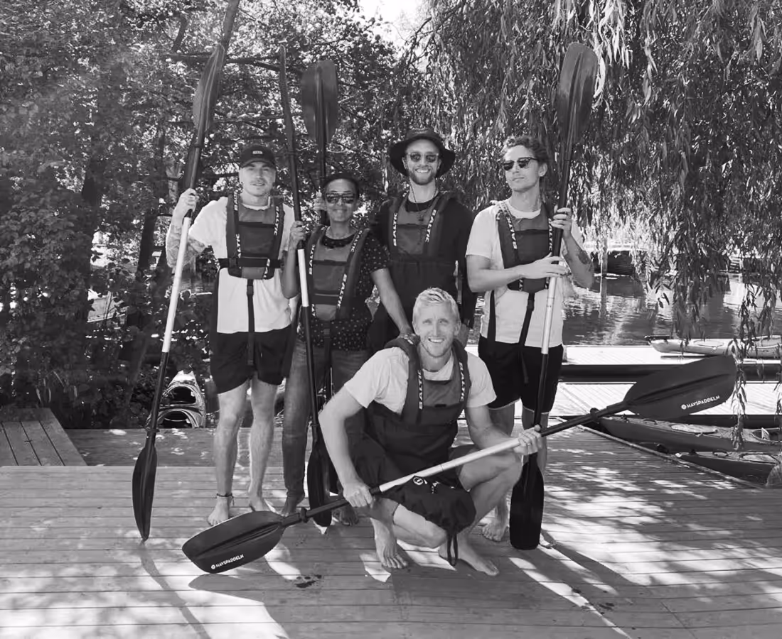 Group of five people on a wooden dock holding kayak paddles, dressed in life vests, with kayaks and trees in the background.