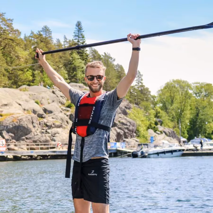 Anton, Co-founder of BookSpot wearing sunglasses, life jacket, and shorts holding a kayak paddle above his head on a sunny day near rocky shoreline and boats.