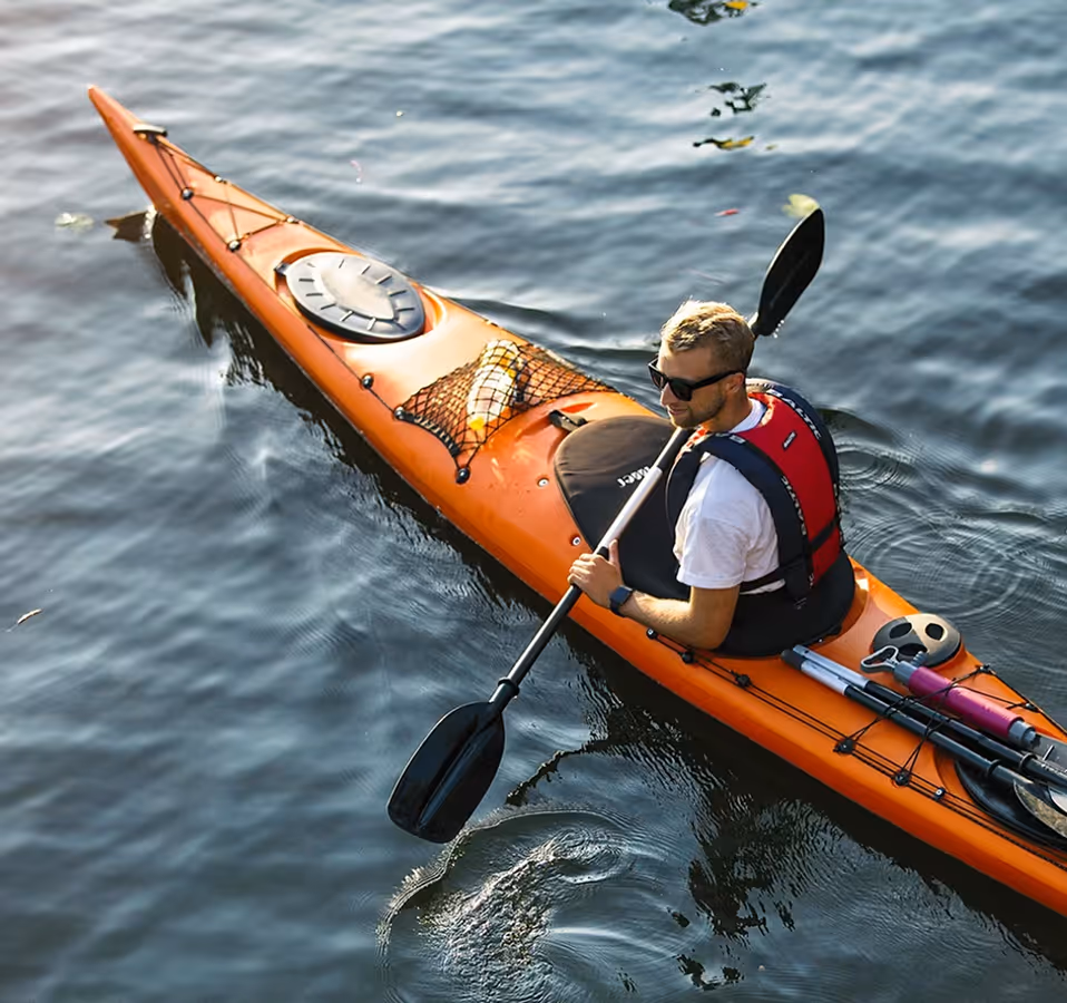 Man wearing sunglasses and a red life vest paddling an orange kayak on calm water.