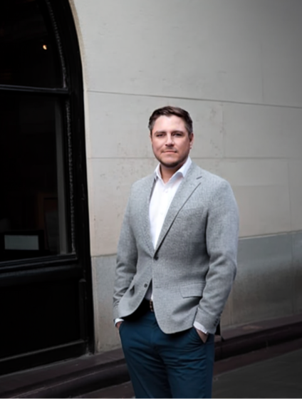 Man wearing a gray blazer and white shirt standing with hands in pockets against a stone wall near a window.