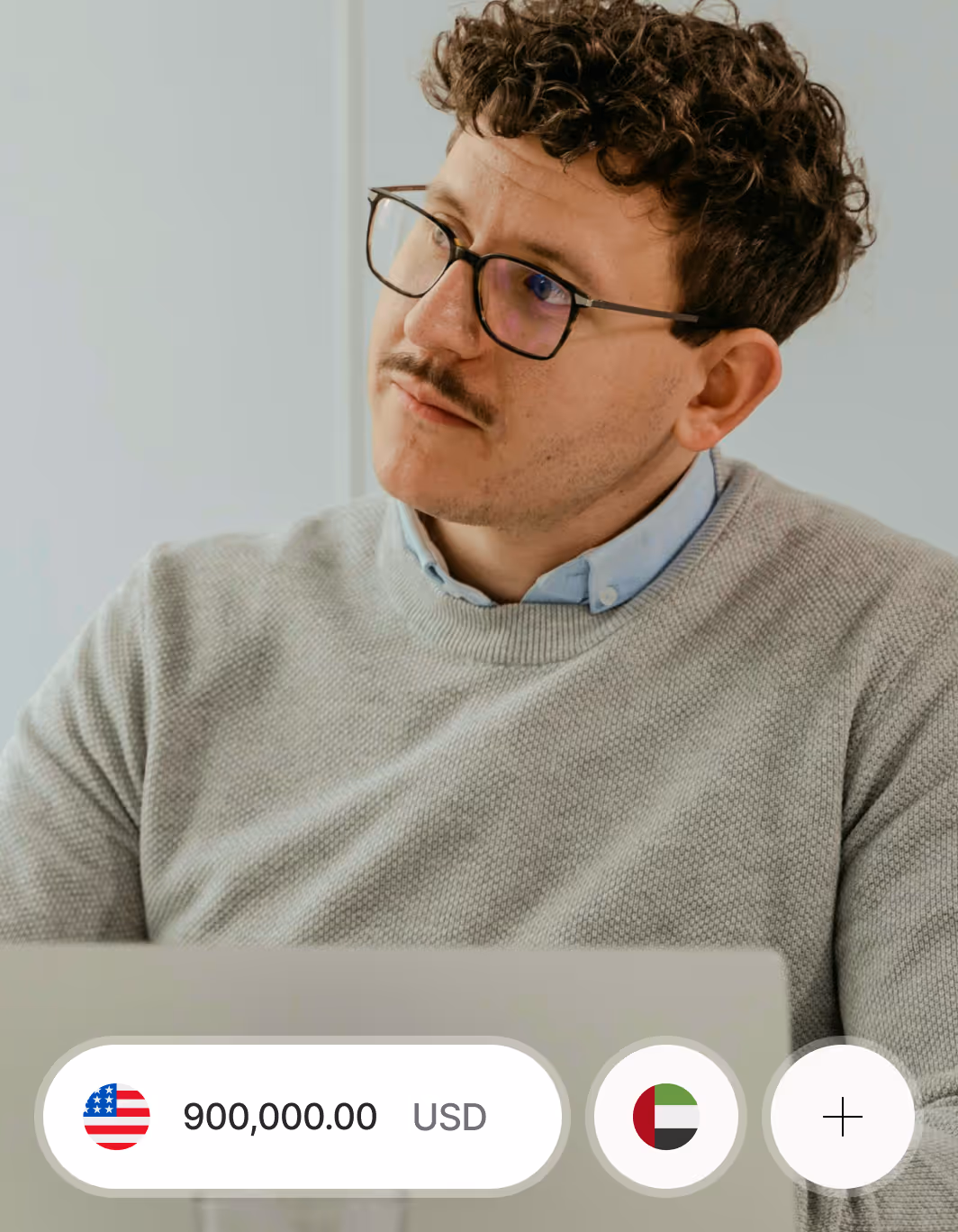Man with glasses and mustache wearing a light gray sweater looking to the side behind a laptop, with currency icons and an amount of 900,000.00 displayed.
