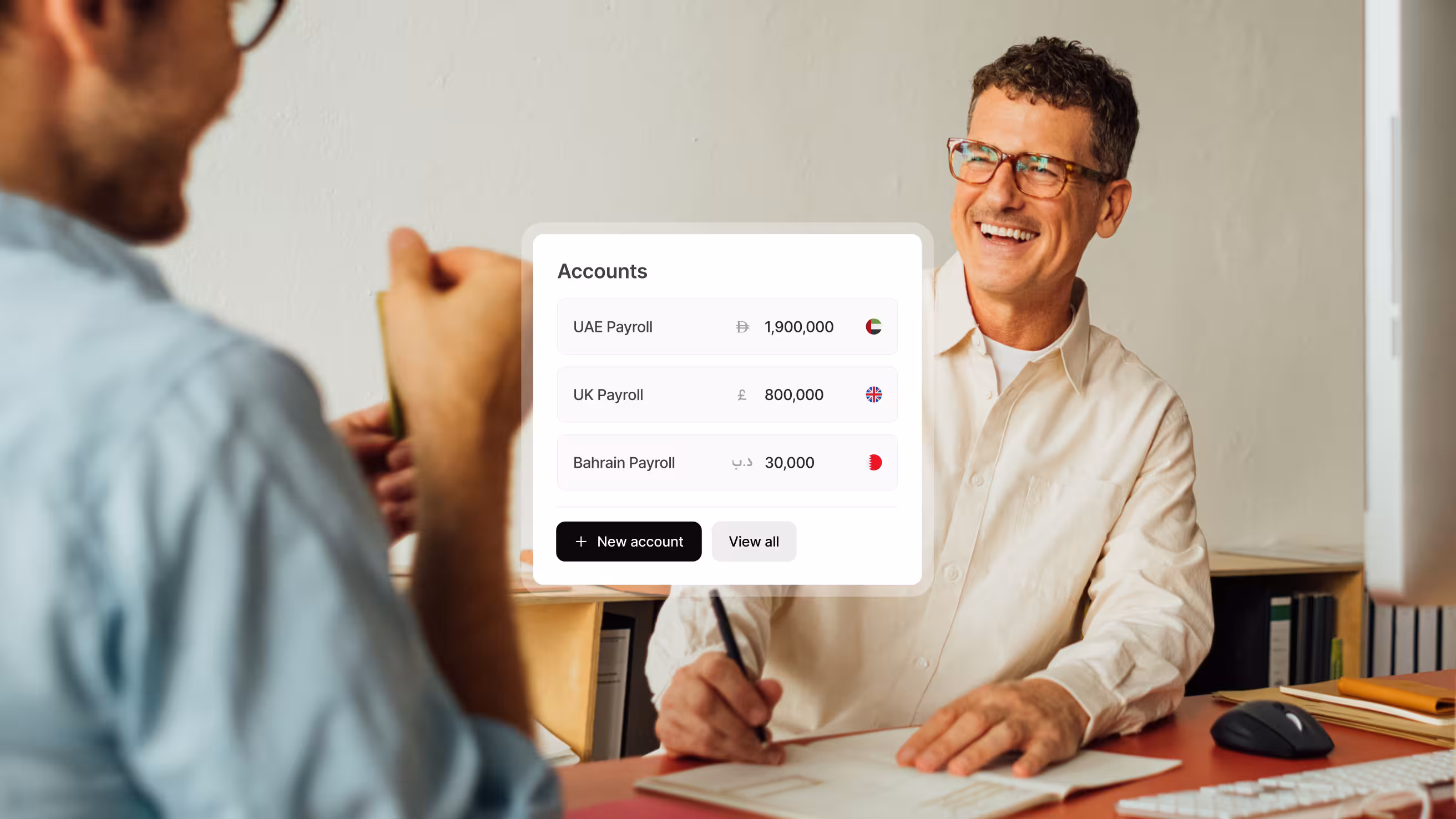 Two people sitting in a bright office with a table between them, showing an overlaid list of payroll accounts for UAE, UK, and Bahrain with their balances.