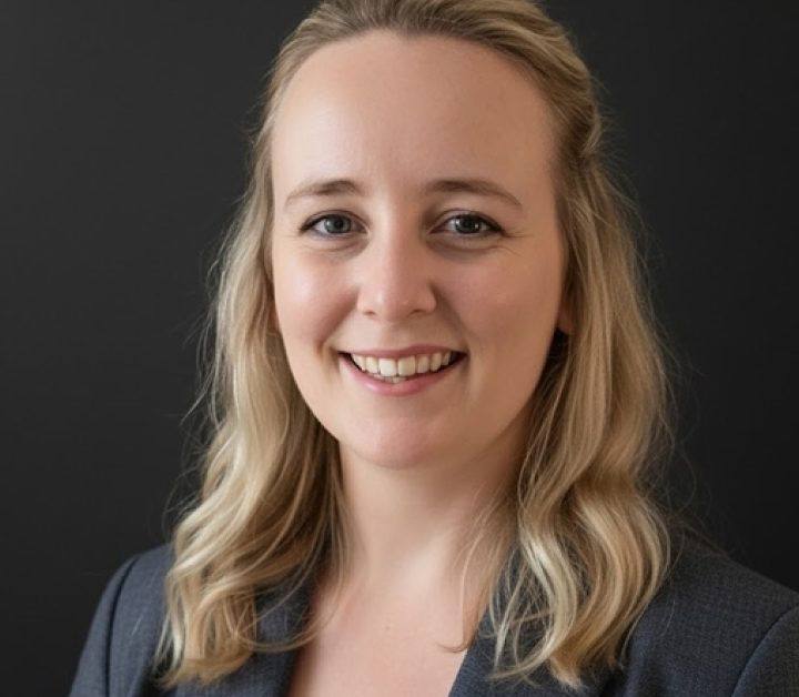 Smiling woman with shoulder-length blonde hair wearing a gray blazer against a dark background.