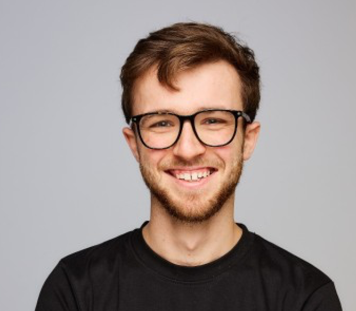 Smiling young man with short brown hair, beard, black glasses, and black shirt against a gray background.