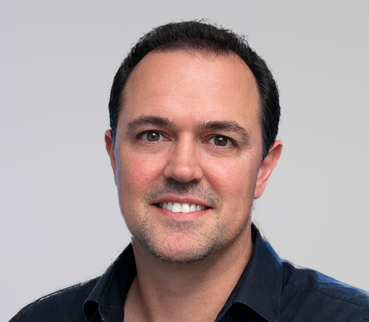 Smiling man with short dark hair wearing a dark navy shirt against a light grey background.