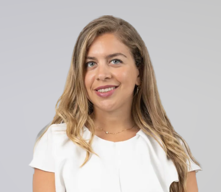 Smiling woman with long blonde hair wearing a white blouse against a light grey background.