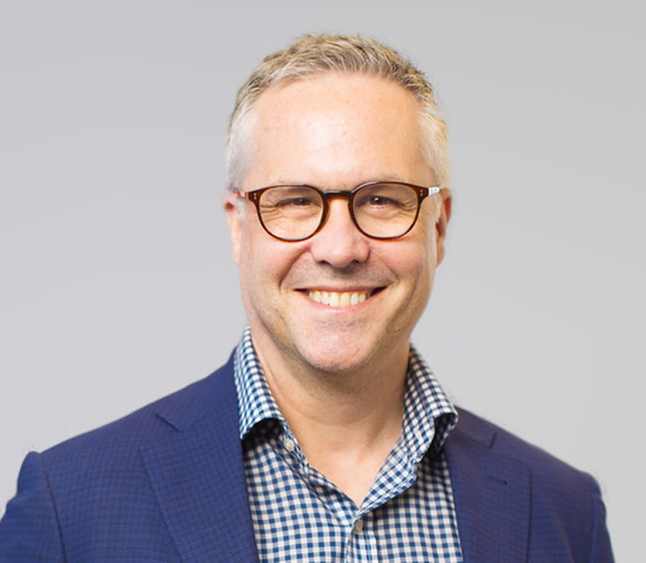 Smiling man with grey hair and tortoiseshell glasses wearing a navy jacket over a checked shirt.