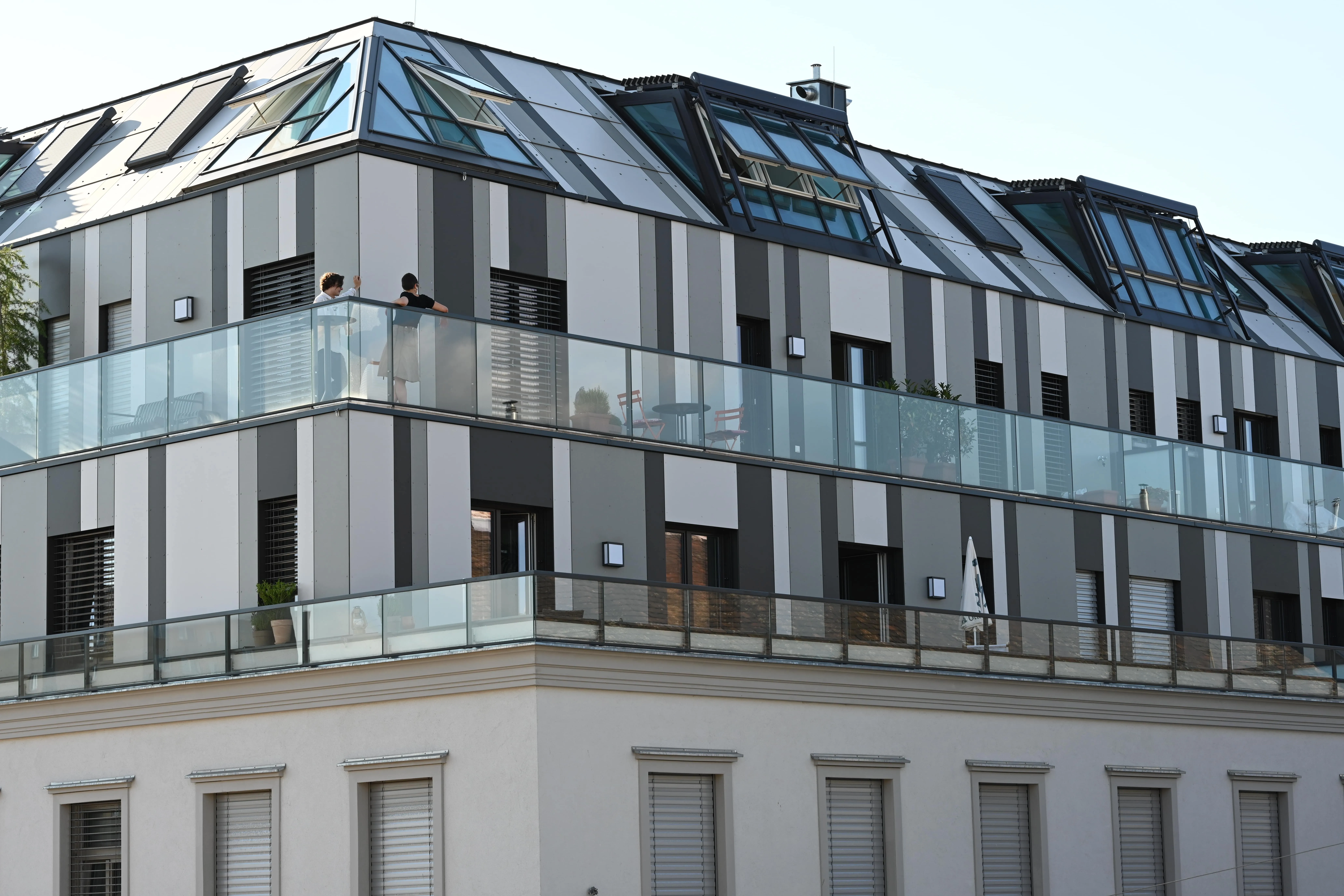 Moderner Wohnkomplex mit einer Terrasse und zwei Personen, die auf den Glasbalustraden stehen.