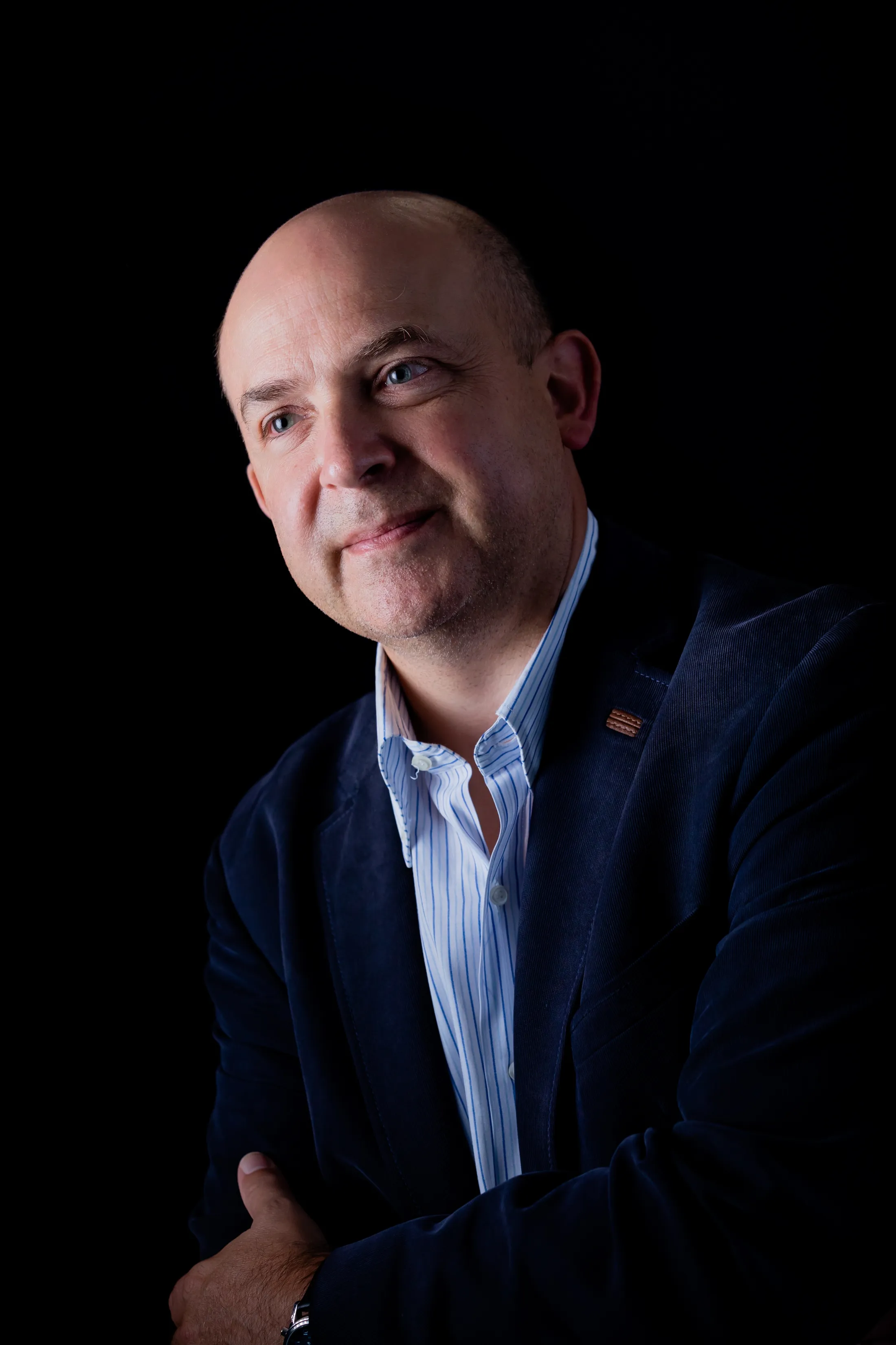 Porträt eines nachdenklich blickenden, glatzköpfigen Mannes in blauem Sakko und gestreiftem Hemd vor schwarzem Hintergrund.