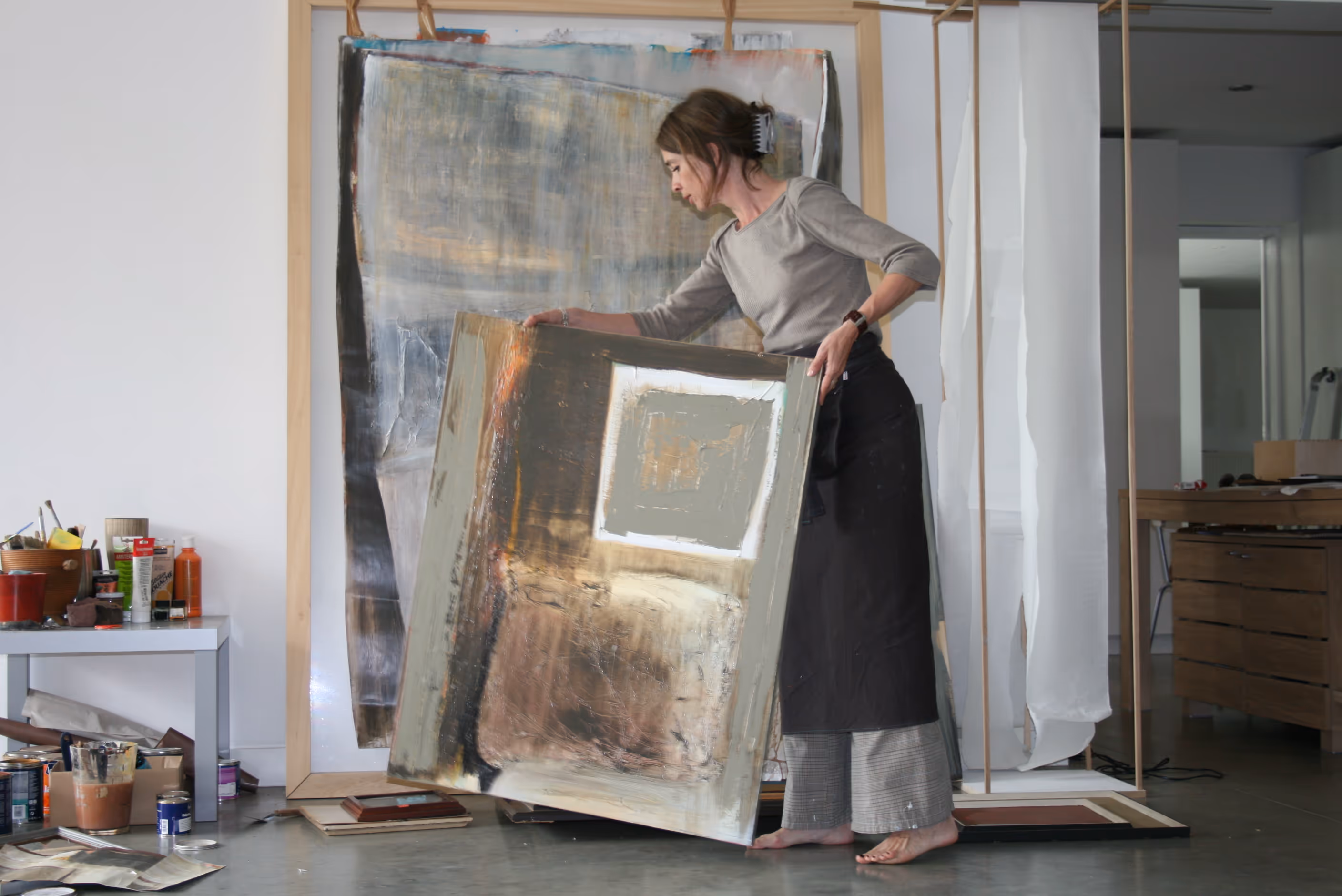 Lies staat op blote voeten en houdt een abstract schilderij vast in een atelier met meer schilderijen en schildermaterialen op de vloer en een tafel.