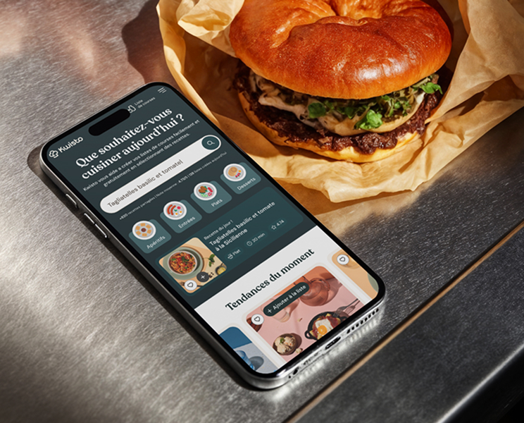 Smartphone affichant une interface d'application de cuisine française à côté d'un burger gourmet emballé sur une surface en métal.
