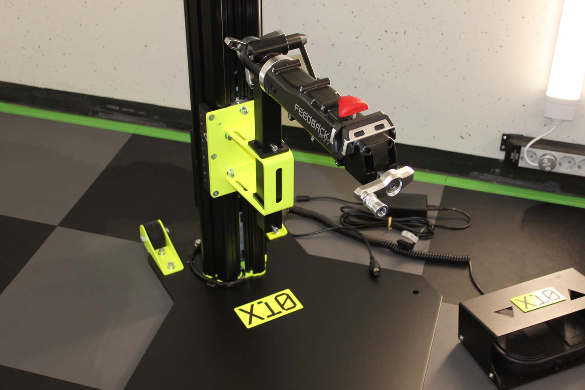 Schwarze und neon-gelbe X10 Fitness-Geräteeinheit mit Griff und Kabeln auf schwarz-grau kariertem Boden.