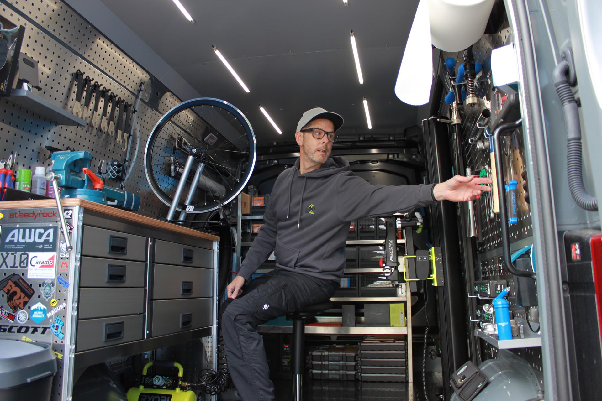 Ein Mann mit Baseballkappe und Brille sitzt auf einem Hocker in einem Werkstattwagen und greift nach Werkzeug an der Wand.