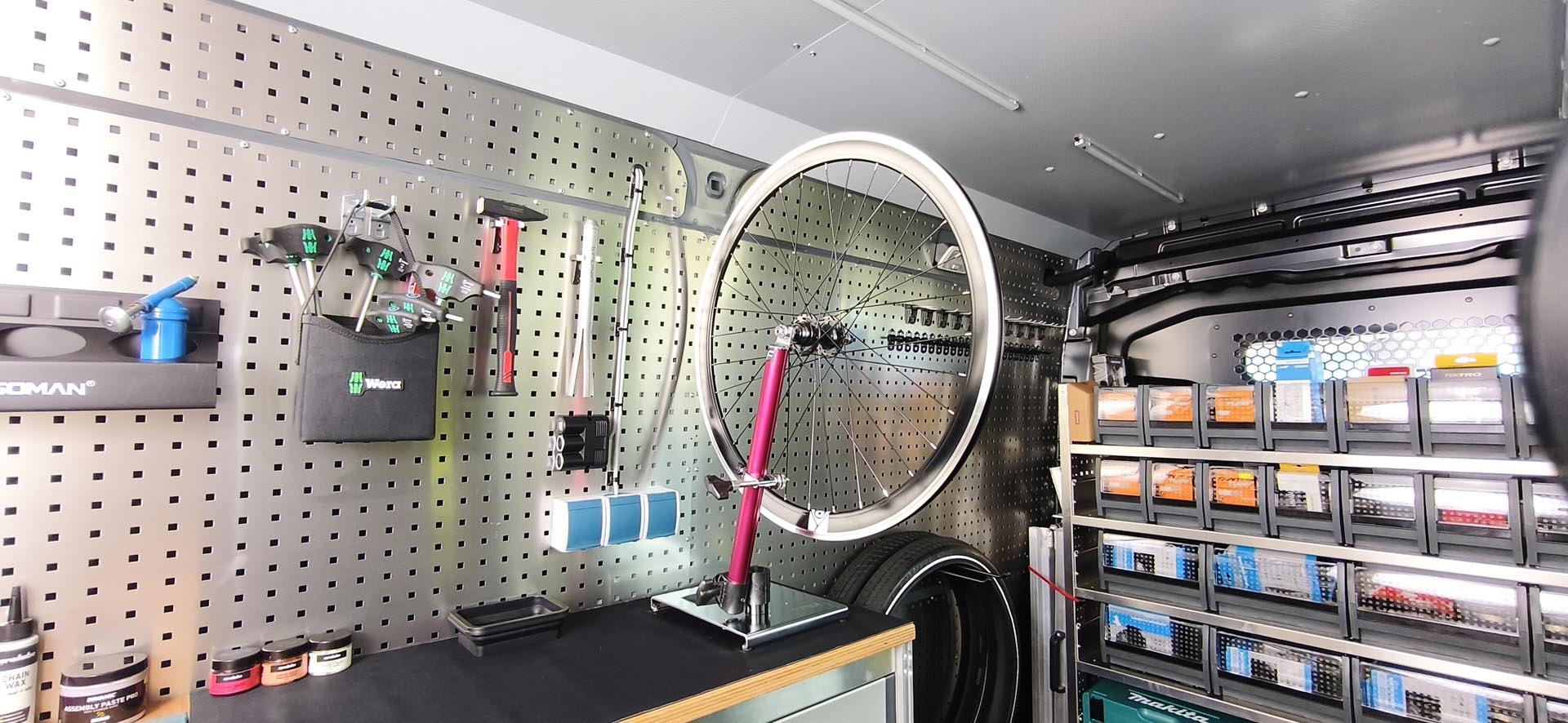 Werkstatt mit einem Fahrradmontageständer, Werkzeug an der Wand und Regalen mit sortierten Kleinteilen.