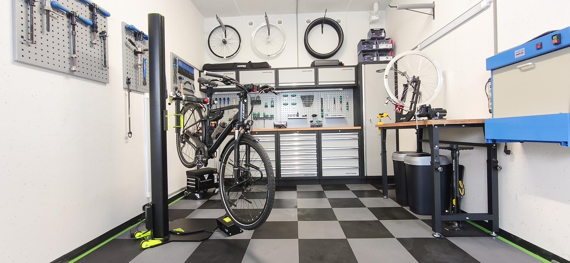 Werkstatt mit einem Fahrrad auf einem Reparaturständer, Werkzeugwänden, Fahrradreifen an der Wand und Arbeitsflächen mit Werkzeugschränken.