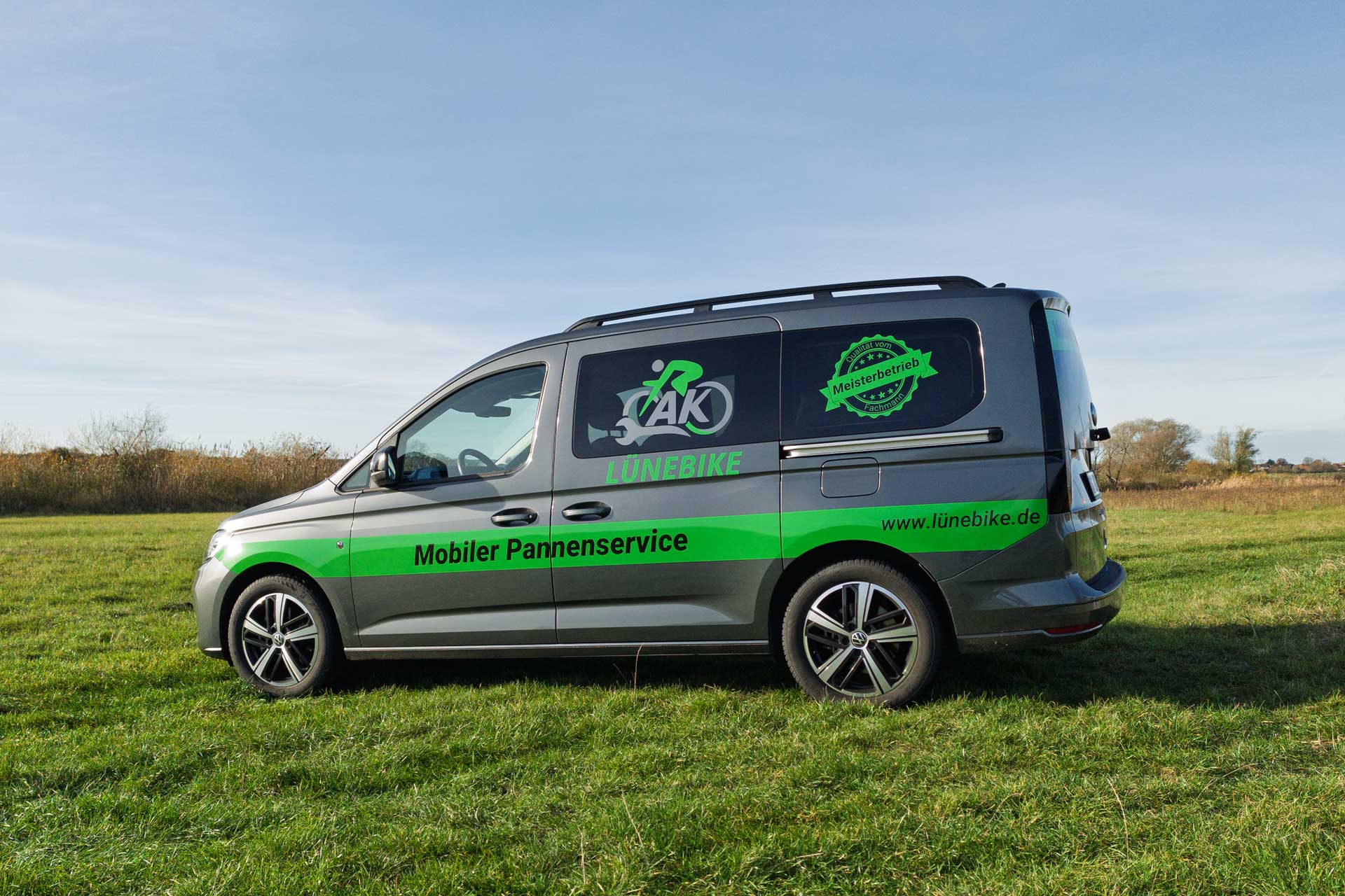 Grauer Lieferwagen mit grüner Beschriftung für mobilen Pannenservice von Lünebike steht auf einer Wiese bei klarem Himmel.