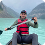 Person wearing a red life jacket and hat paddling a kayak on a clear blue lake surrounded by mountains.