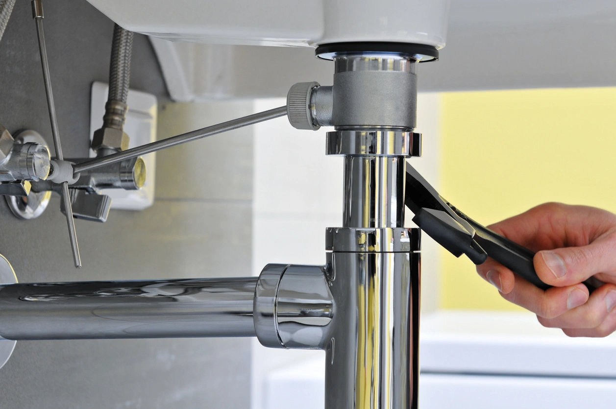 A person using a wrench to adjust a shiny chrome pipe under a sink. 