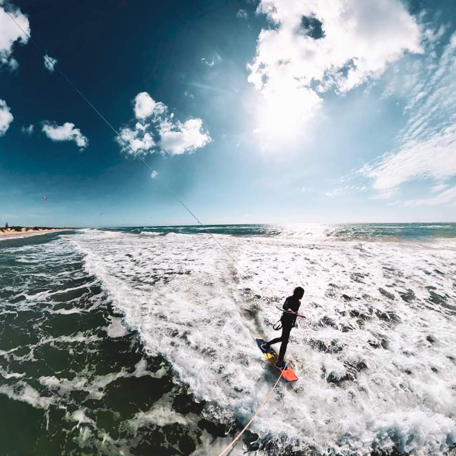 Kitesurfer riding waves along the scenic Sicilian coastline near Floripa Surf and Yoga House, captured in a wide-angle shot under blue skies