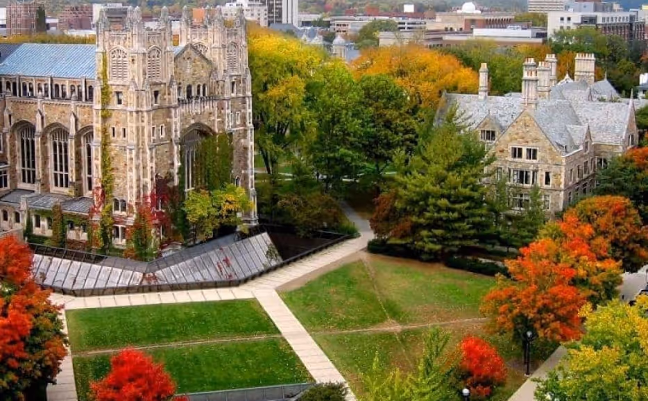 Aerial view of a university campus in autumn showing gothic-style stone buildings surrounded by green lawns and trees with red, orange, and yellow foliage.