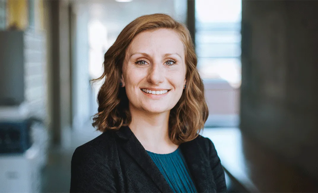 Porträt einer lächelnden Geschäftsfrau mit roten Haaren, die einen blauen Pullover und ein schwarzes Sakko trägt.