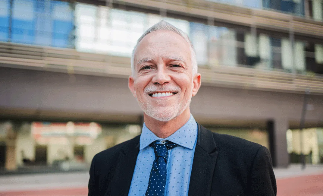 Lächelnder älterer Geschäftsmann mit grauen Haaren und Bart in Anzug und Krawatte vor modernem Gebäude.