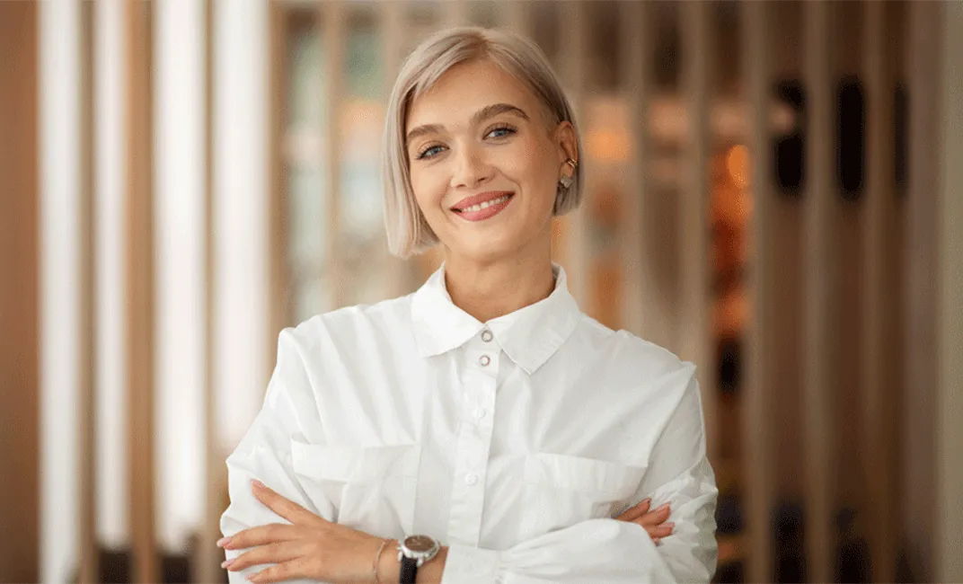 Lächelnde Frau mit blonden kurzen Haaren, die weiße Bluse trägt und die Arme verschränkt hat.