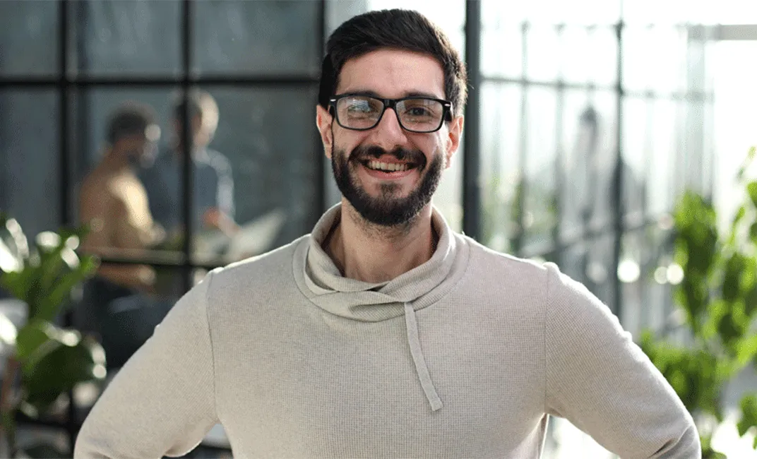 Ein lächelnder Mann mit schwarzer Brille und Bart trägt einen hellen Pullover und steht vor einem Büro mit Glaswänden.