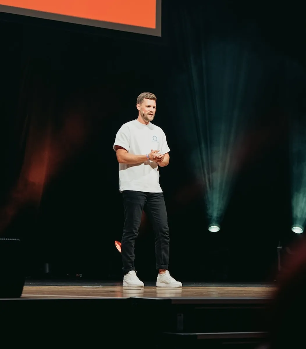 Ein Mann in weißem T-Shirt und schwarzer Hose hält eine Rede auf einer Bühne mit Bühnenlicht im Hintergrund.