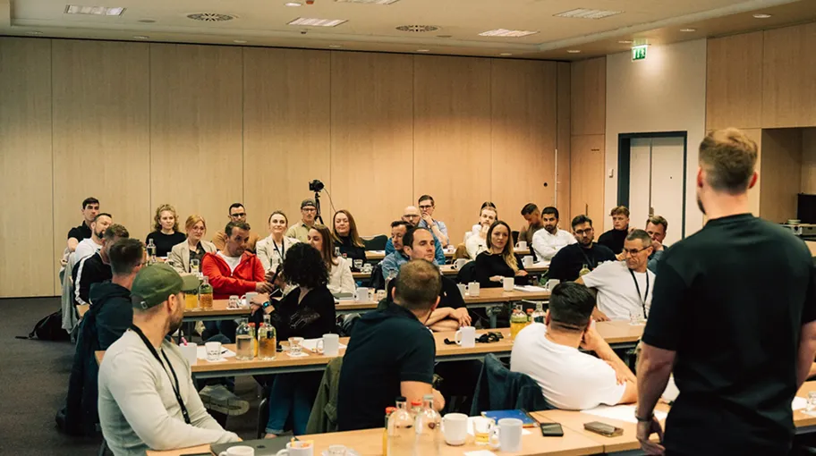 Gruppe von Erwachsenen sitzt in einem Klassenzimmer und hört aufmerksam einem stehenden männlichen Redner zu.