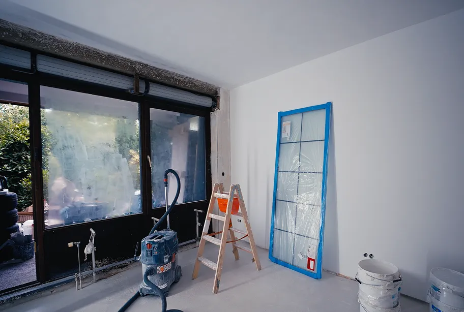 Innenraum in Renovierung mit angelehnter verpackter Glastür, Holzleiter, Staubsauger und Eimern auf dem Boden vor großen Fenstern.