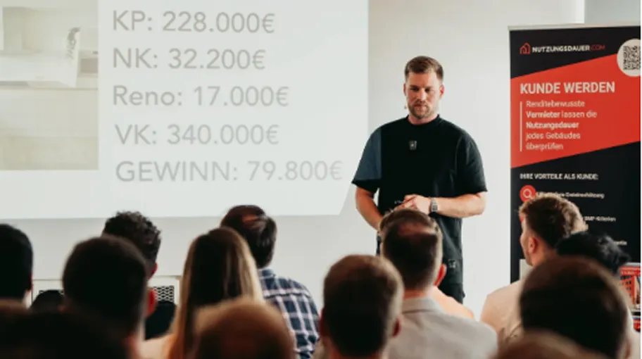 Ein Mann hält eine Präsentation vor sitzendem Publikum mit Zahlen zu Kosten und Gewinn auf einer Projektion und einem Werbebanner im Hintergrund.