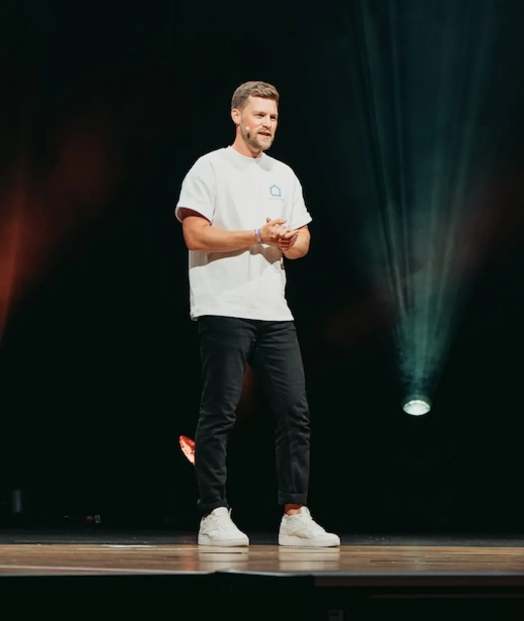 Mann mit weißen Turnschuhen, schwarzer Hose und weißem T-Shirt spricht auf einer Bühne mit dunklem Hintergrund und Bühnenlicht.