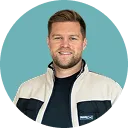 Porträt eines lächelnden Mannes mit kurzem Haar, der eine blaue Jacke trägt vor einem einfarbigen türkisfarbenen Hintergrund.