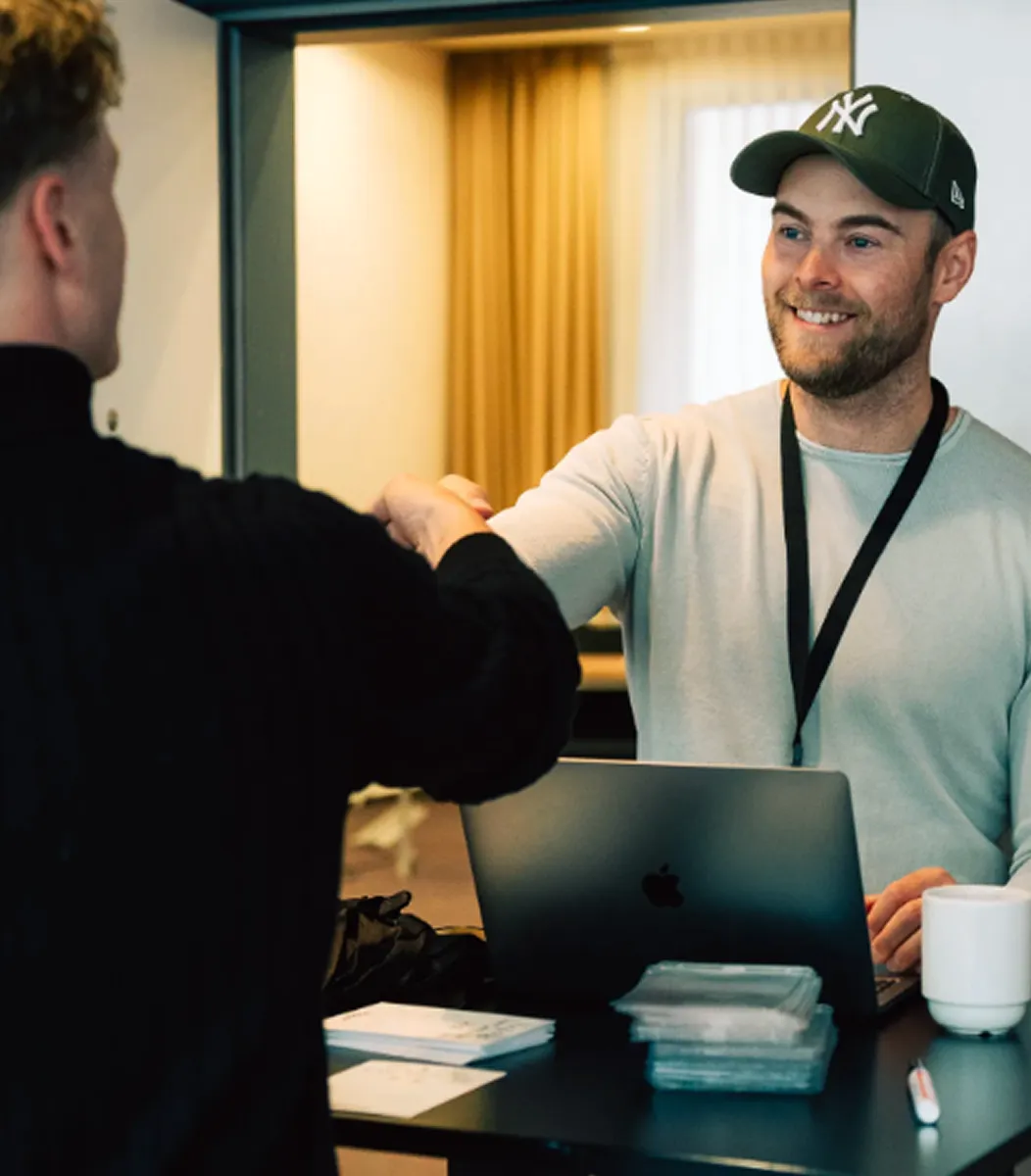 Zwei Männer begrüßen sich per Ellbogenstoß an einem Tisch mit Laptop, Kaffeetasse und Dokumenten.