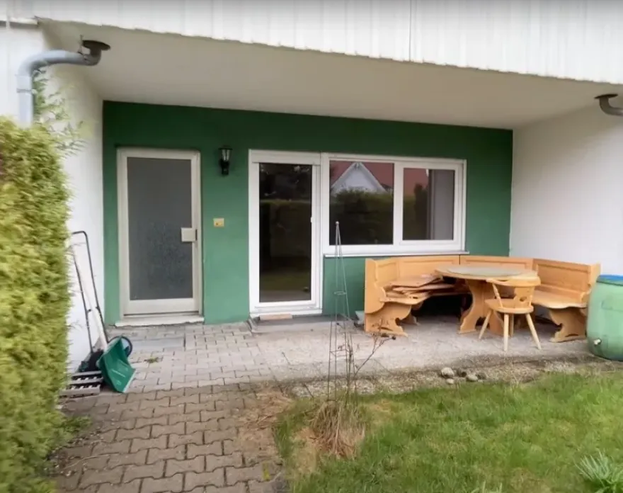 Terrasse mit einer grünen Wand, zwei Türen, einem Fenster und einer hellen Holzecke mit Tisch und Stühlen neben einem grünen Regentonne.